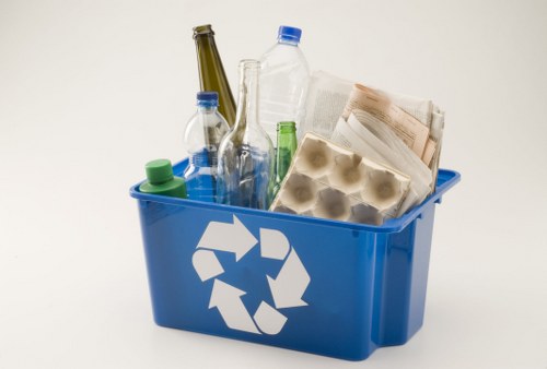 A blue plastic recycling bin with a white recycling symbol on the front, filled with various waste items including clear and brown glass bottles, plastic water bottles with blue and green caps, a cardboard egg carton, and crumpled paper or newspaper. The bin is placed on a plain white surface with some waste materials leaning and slightly protruding from the top. The setting appears to be a clean, indoor environment, possibly a work or storage area in London, as part of a commercial waste removal service based in Earls Court, focused on rubbish collection and disposal.