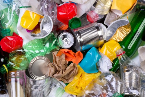A close-up view of a mixed pile of recyclable waste materials, including crushed aluminium cans with reflective metallic surfaces, crumpled paper and brown card, and various plastic bottles and containers in different colours such as green, yellow, red, and transparent. The plastic bottles feature screw caps attached or removed, and the materials are loosely arranged on a surface, typical of waste collected for rubbish removal services. The background shows more scattered plastic and metal waste, indicating a cluttered collection point or skip load. The textures range from smooth and shiny metallic cans to crinkled paper and soft plastic. This image visually represents household or commercial waste that a company like Commercial Waste Removal Earls Court would handle, emphasizing the importance of proper rubbish disposal and management in a local London context near Earls Court.