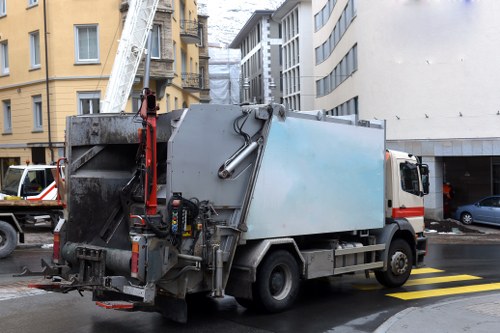 A large commercial waste collection truck parked on a city street verge between multi-story buildings, with a silver metallic cargo container and a lifting mechanism at the rear, used for rubbish collection and disposal; the truck is positioned near a pedestrian crossing with yellow and black painted stripes, and the surrounding environment includes residential and commercial structures with balconies, windows, and some construction or scaffolding visible in the background. The scene is set during daytime with natural light, and the truck’s exterior appears slightly weathered, typical of heavy-duty waste management vehicles used by Commercial Waste Removal Earls Court for rubbish clearance services in the area.
