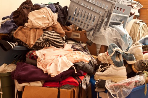 A large pile of assorted domestic laundry and household textiles, including clothes, towels, and fabric items, situated on a wooden surface in an indoor setting. The clothes are varied in colour, with some light and some dark, including beige, brown, black, red, and cream tones, and are a mix of different textures, from smooth cotton to thicker, knit materials. Several items are draped over or partially covering a cardboard box, with some garments folded and others loosely piled. In the background, there are plastic laundry baskets, one of which is tipped slightly to the side, revealing additional clothing inside, and a stack of shoes, including sneakers and sandals, placed at the edge of the heap. The environment appears to be a storage or utility room with warm ambient lighting, indicating a typical household space in the vicinity of Earls Court, London, where commercial waste removal services from Commercial Waste Removal Earls Court are commonly engaged for clearing such clutter.