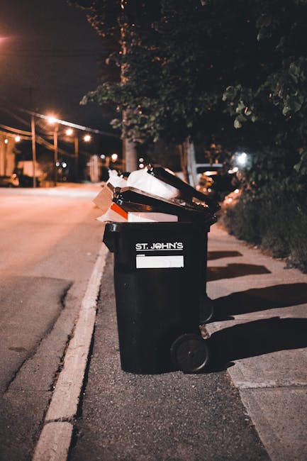 A small, single-axle utility cart loaded with discarded household items and waste materials, positioned on an urban street. The cart contains various large, white cardboard boxes, some open and partia