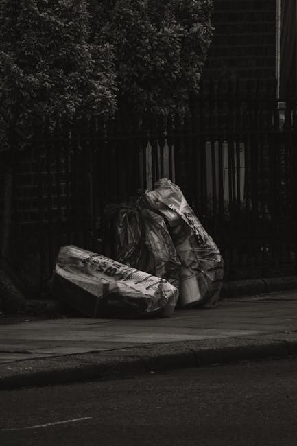 Two large black plastic rubbish bags, filled and knotted, are placed on the pavement next to a dark metal fence with vertical bars, behind which there is dense foliage of trees or bushes. The bags app